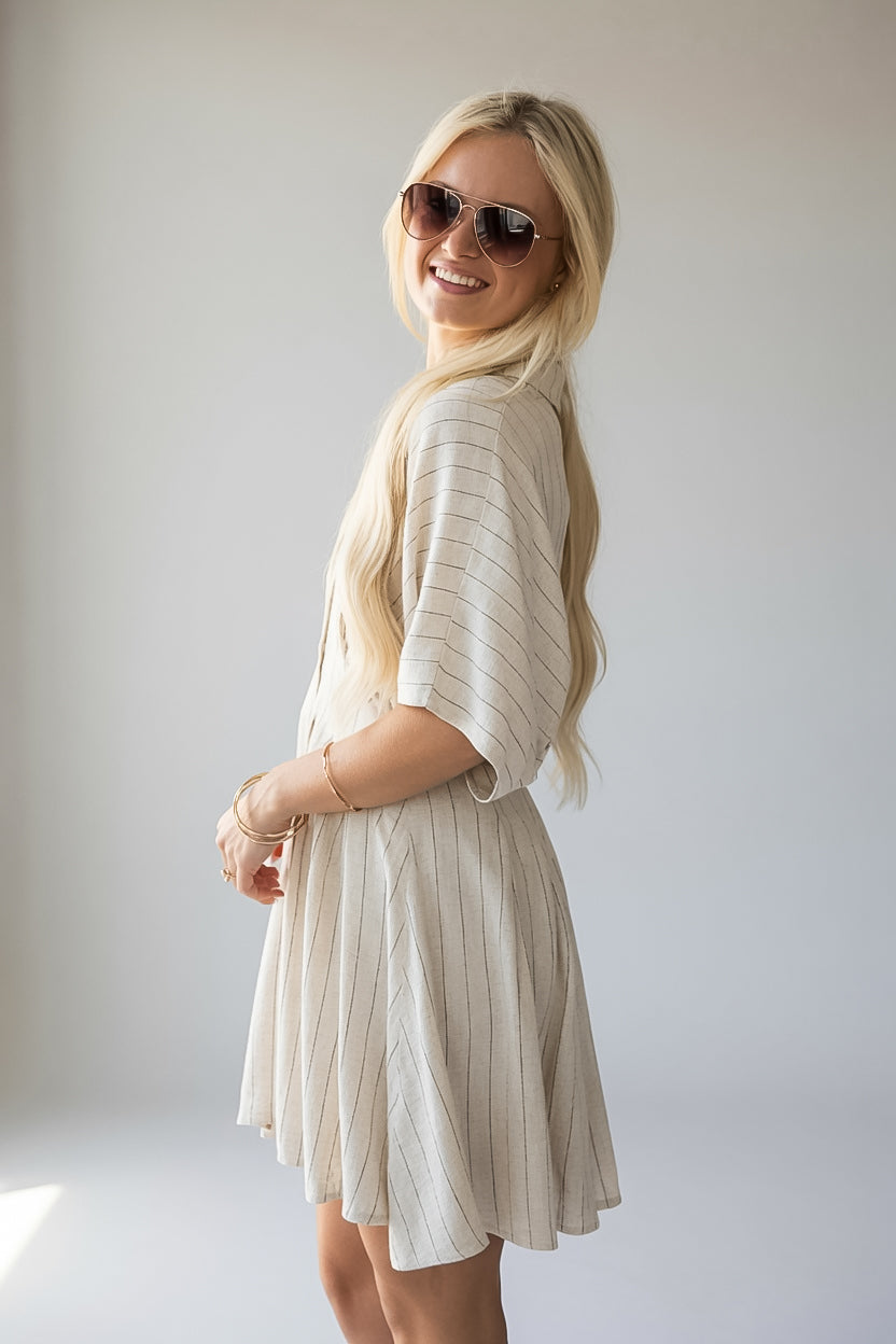 A blonde woman wearing a cream-colored pleated short-sleeve dress and sunglasses, posing in a studio setting against a light gray background.