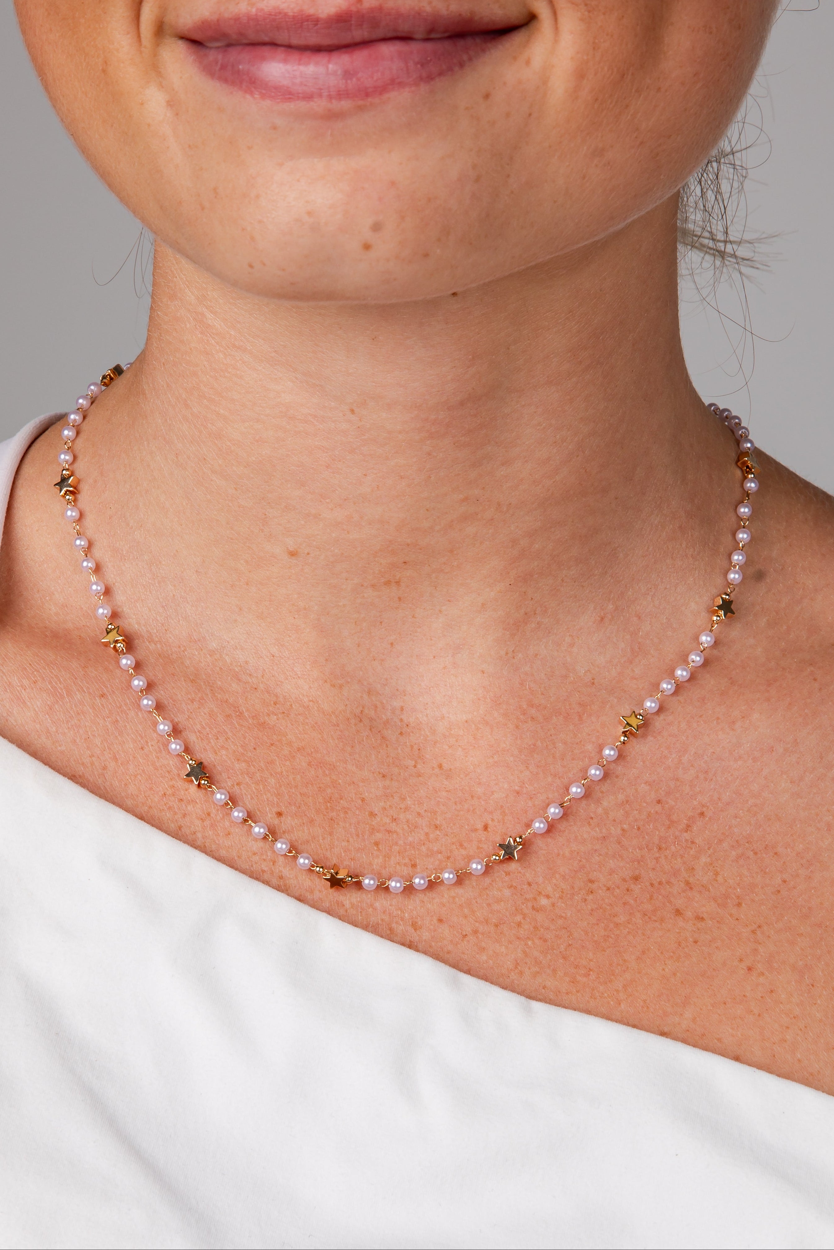 A close-up of a model wearing a delicate necklace made of small pale pearls and gold star-shaped accents, styled with a white off-the-shoulder top against a neutral gray background.