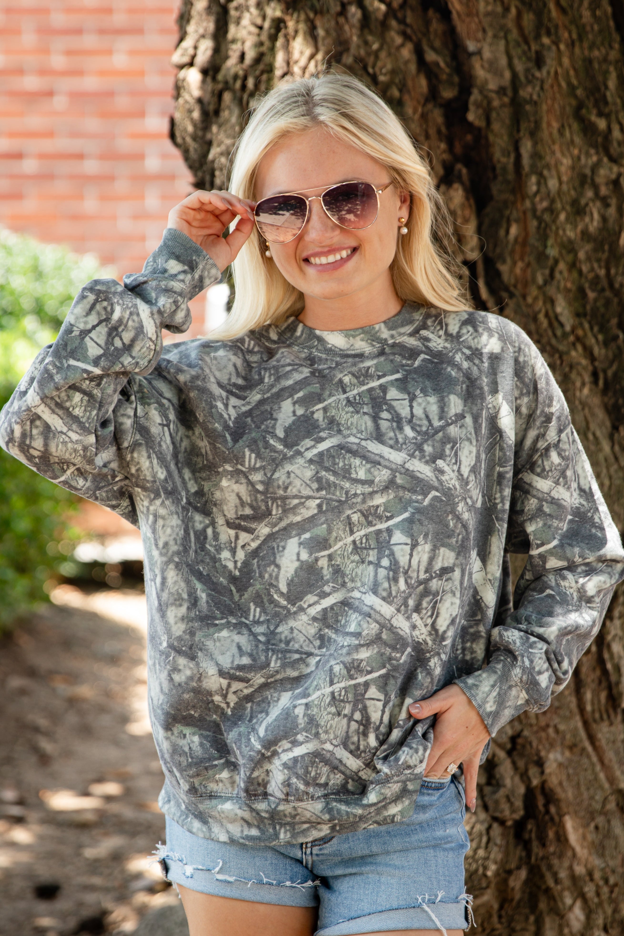 A woman wearing a long-sleeve crewneck sweatshirt with a gray and brown camouflage tree-bark pattern, paired with denim shorts, standing outdoors next to a large tree trunk.