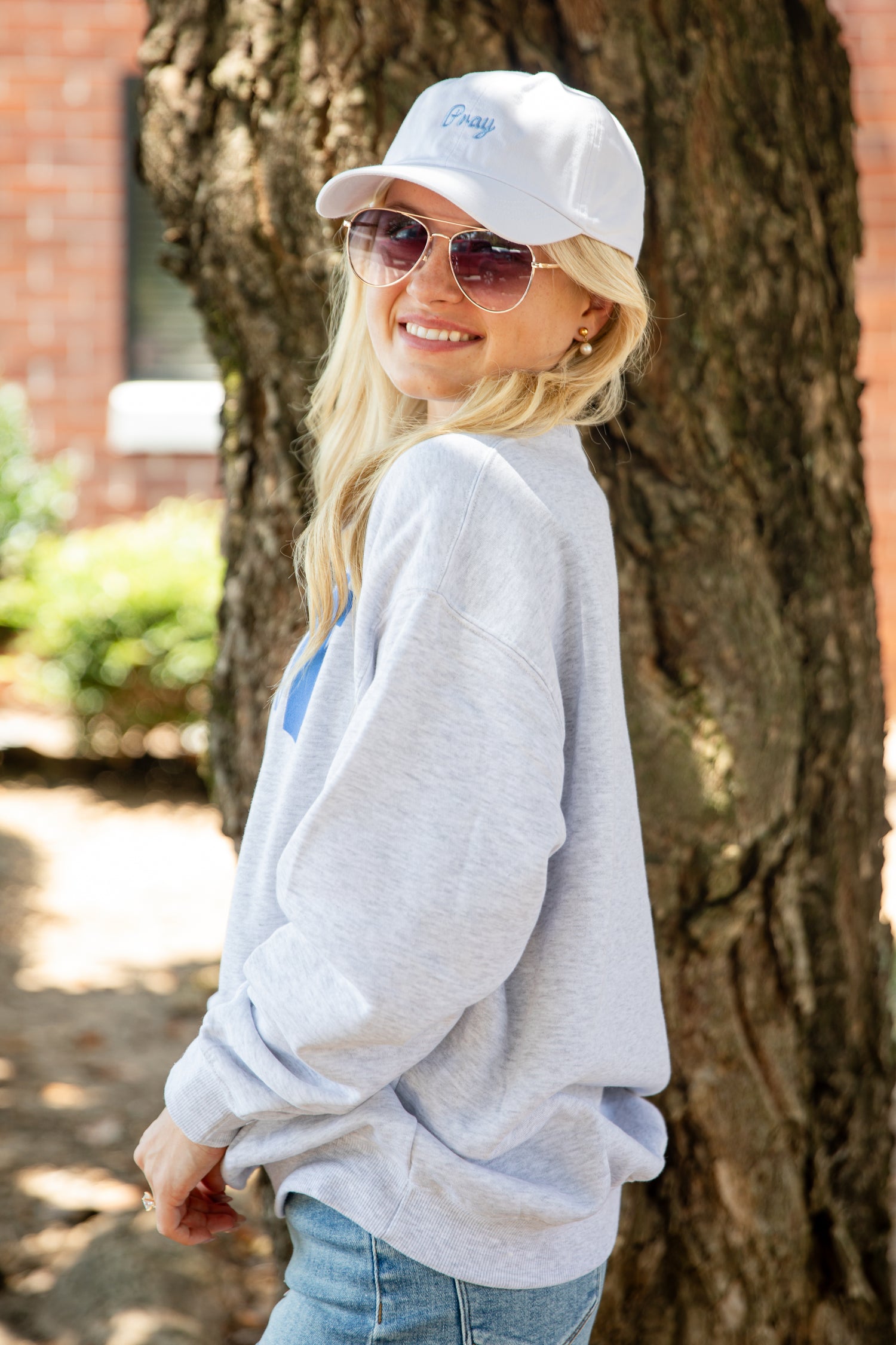 A woman wearing a light heather gray sweatshirt, blue jeans, a white baseball cap, and sunglasses stands outdoors next to a large tree trunk.
