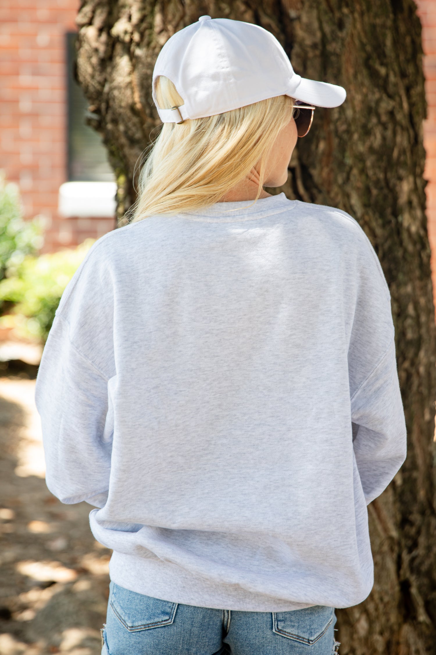 A woman viewed from behind wearing a light heather gray crewneck sweatshirt, a white baseball cap, and blue jeans, standing outdoors next to a tree trunk.