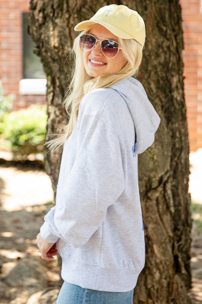 A woman wearing a light gray heathered hoodie, a pale yellow baseball cap, and sunglasses stands outdoors next to a tree trunk.
