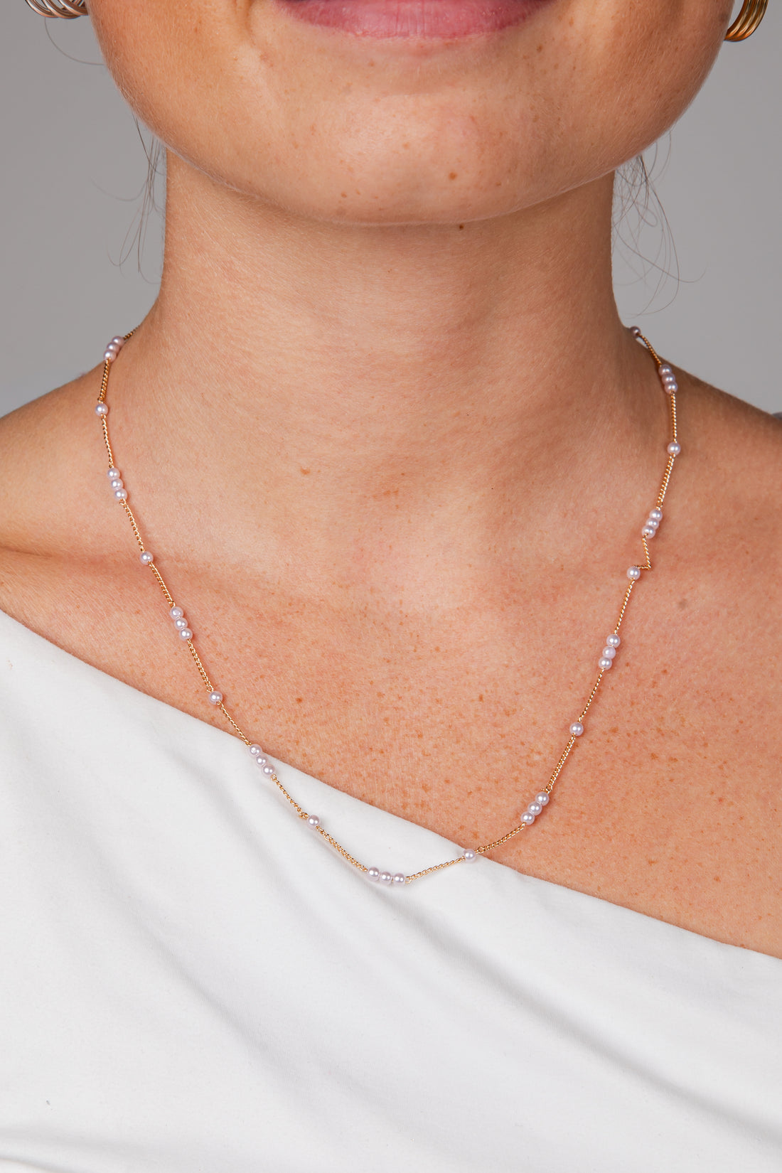 A close-up of a model wearing a delicate rose gold chain necklace with small white pearl-like beads spaced along the chain, paired with a white off-the-shoulder top.