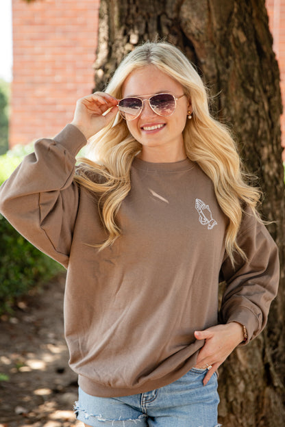 A woman wearing a brown crewneck sweatshirt with a small white graphic on the left chest, paired with denim shorts, posing outdoors in front of a tree.