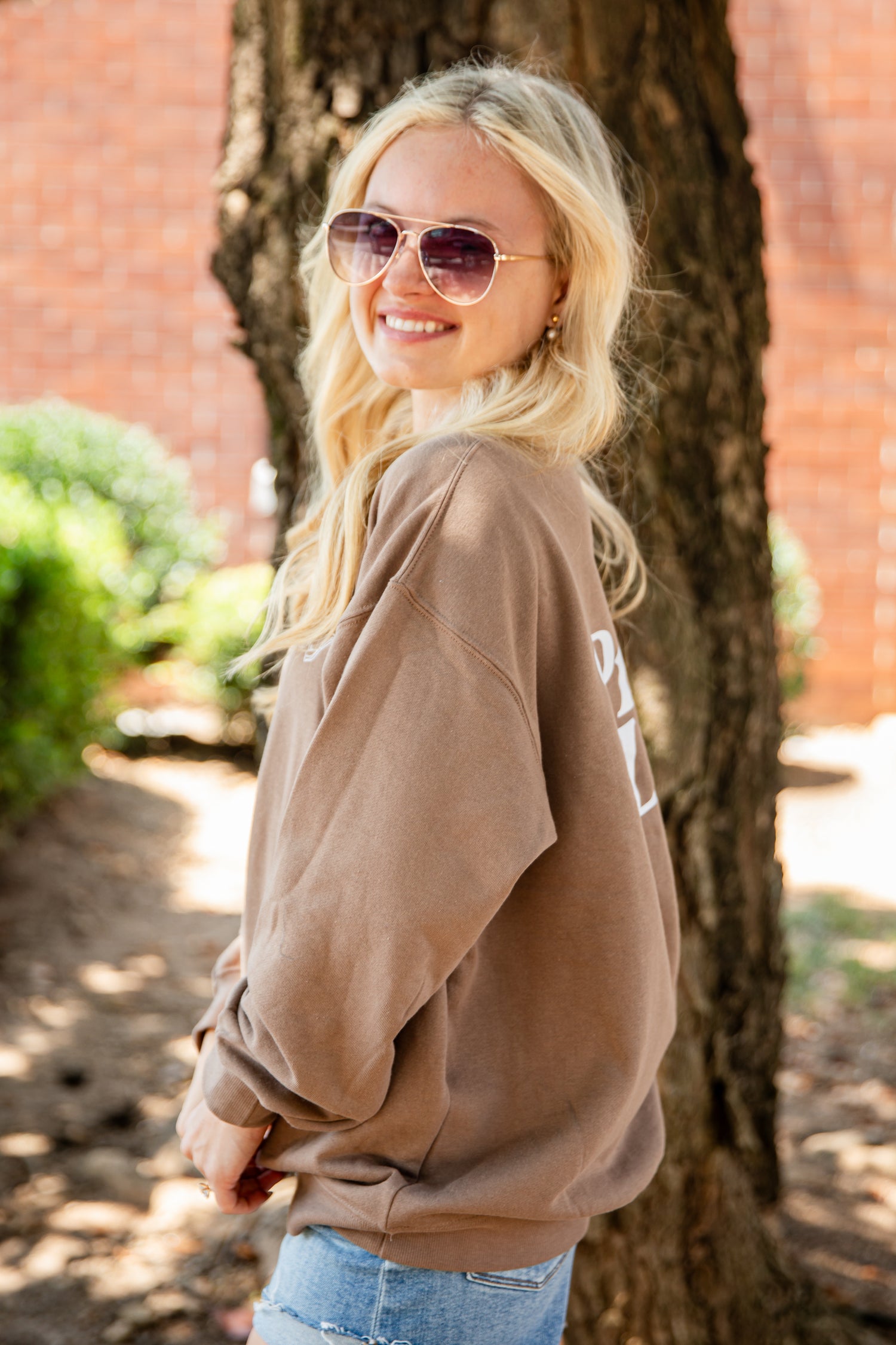 A blonde woman wearing aviator sunglasses and a brown oversized sweatshirt with white lettering, standing outdoors next to a tree trunk.