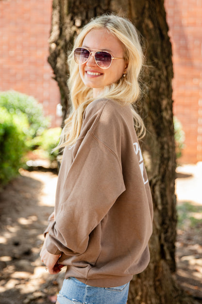 A blonde woman wearing aviator sunglasses and a brown oversized sweatshirt with white lettering, standing outdoors next to a tree trunk.