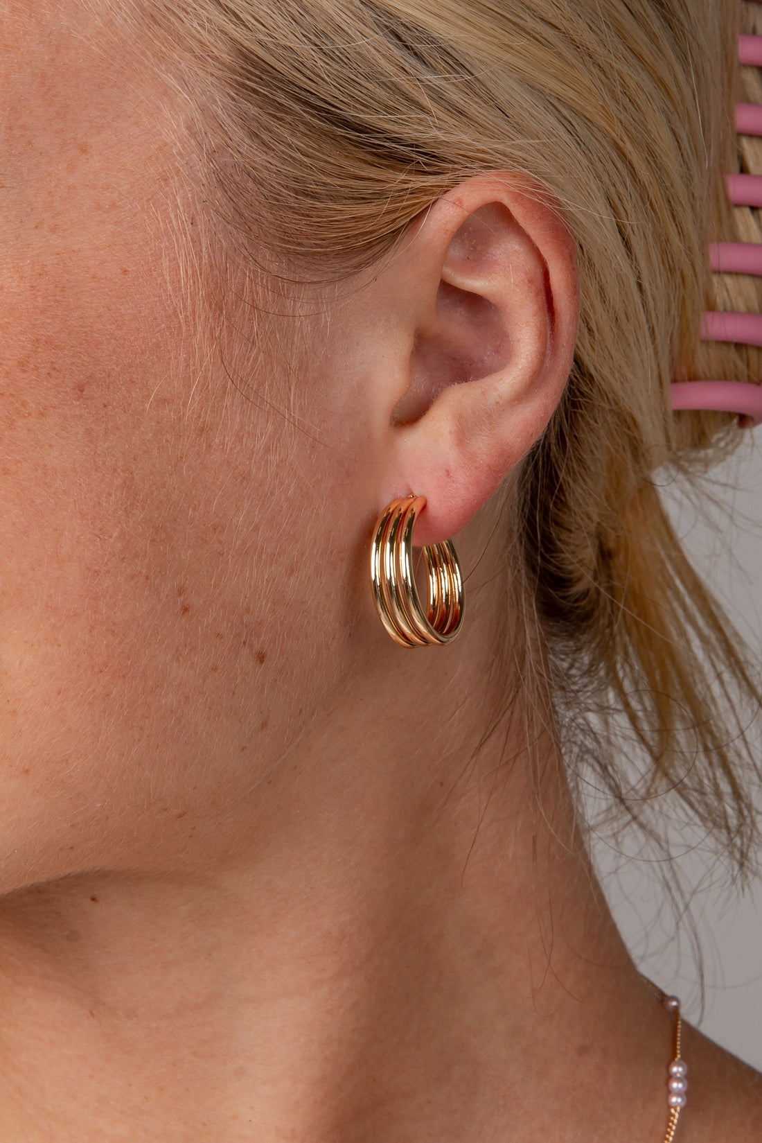 A close-up side profile of a woman with blonde hair in a pink claw clip, wearing a pair of gold triple-hoop earrings and a delicate pearl necklace against a neutral gray background.
