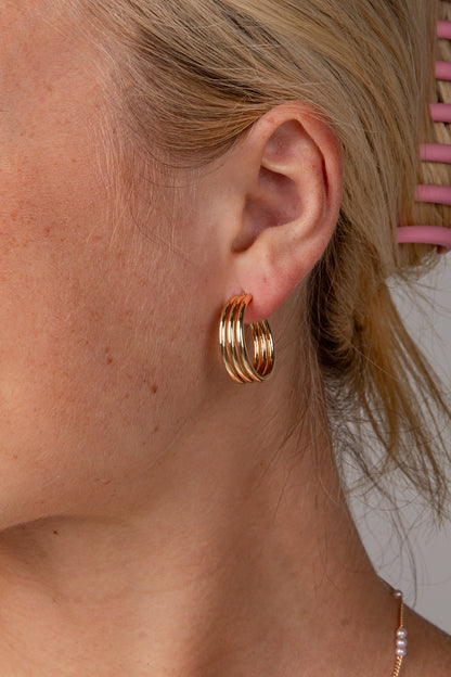 A close-up side profile of a woman with blonde hair in a pink claw clip, wearing a pair of gold triple-hoop earrings and a delicate pearl necklace against a neutral gray background.