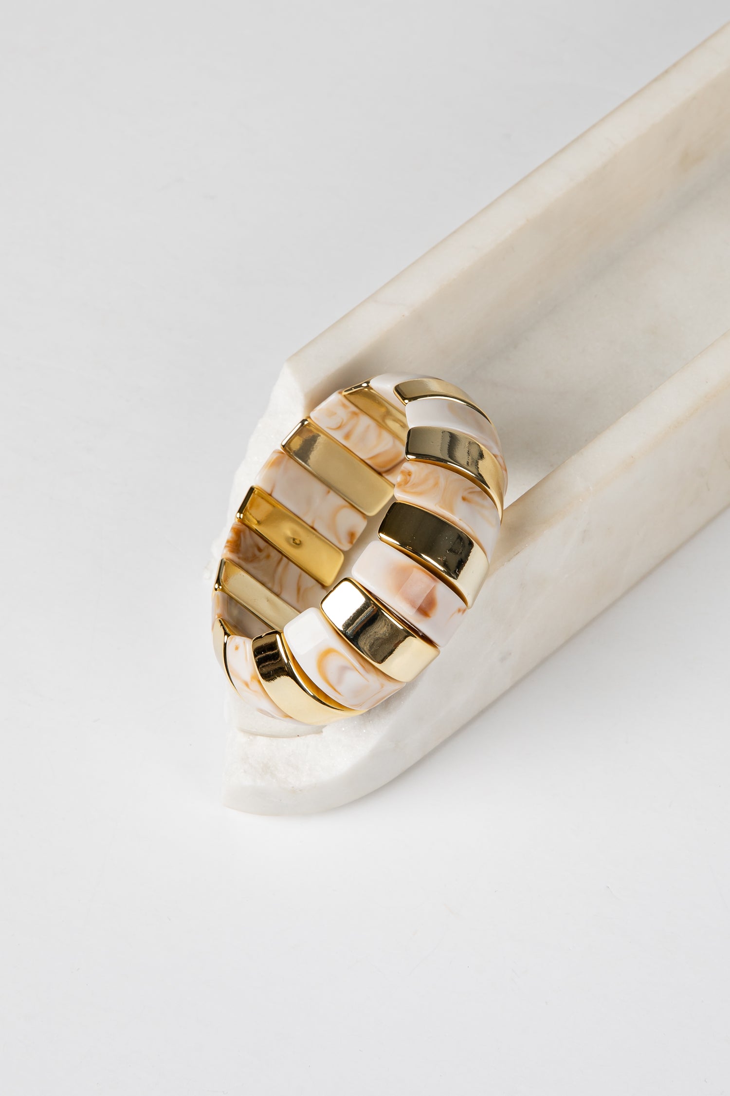 A wide statement ring with alternating polished gold and iridescent white mother-of-pearl segments, displayed on a white marble stand against a clean white background.