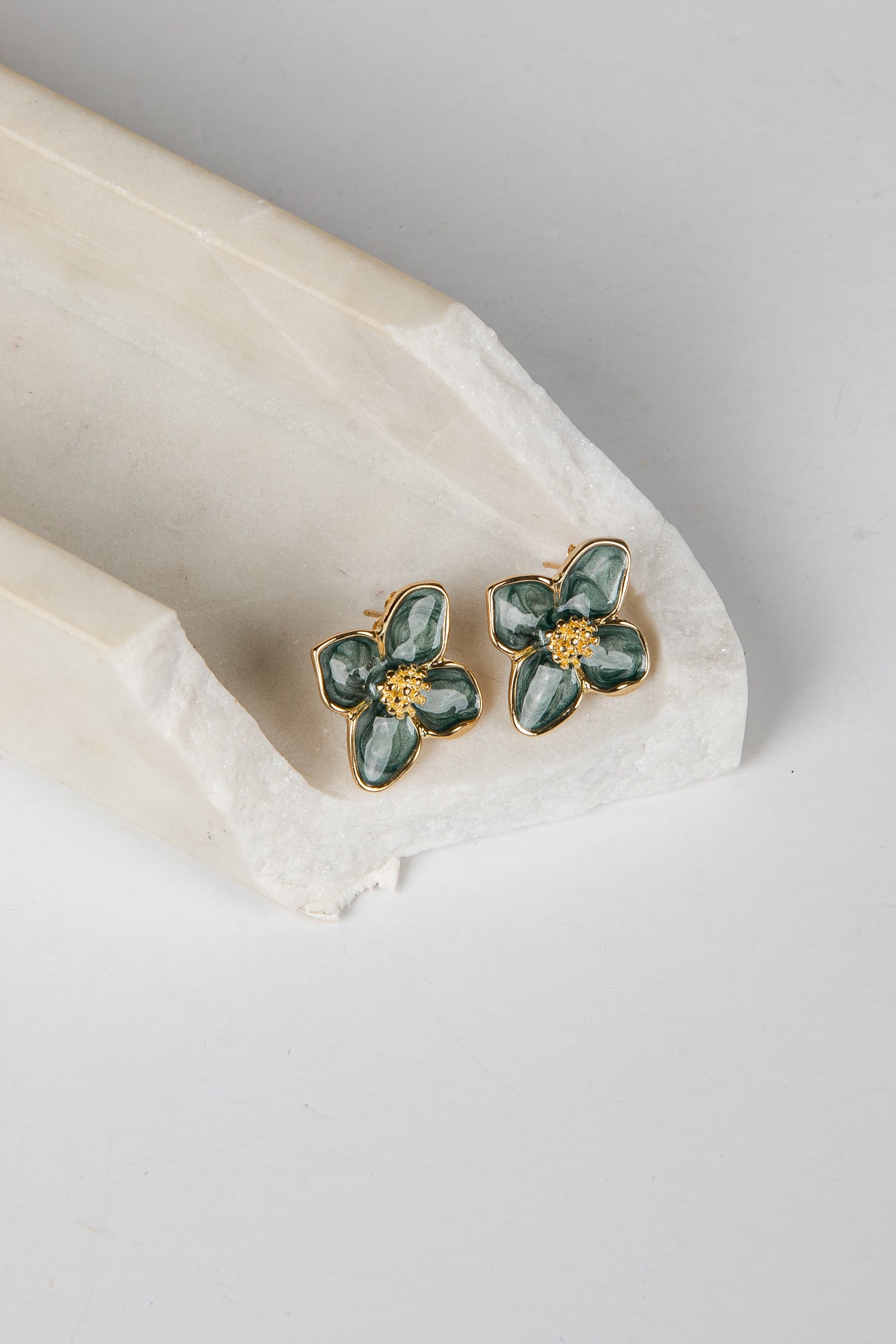A pair of gold flower-shaped stud earrings with green enamel petals, resting on a white marble tray against a light gray background.