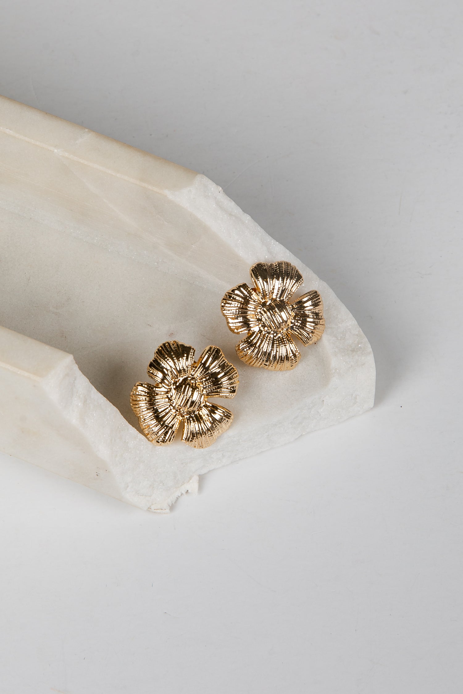 A pair of textured gold flower-shaped stud earrings resting on a white marble tray against a light gray background.