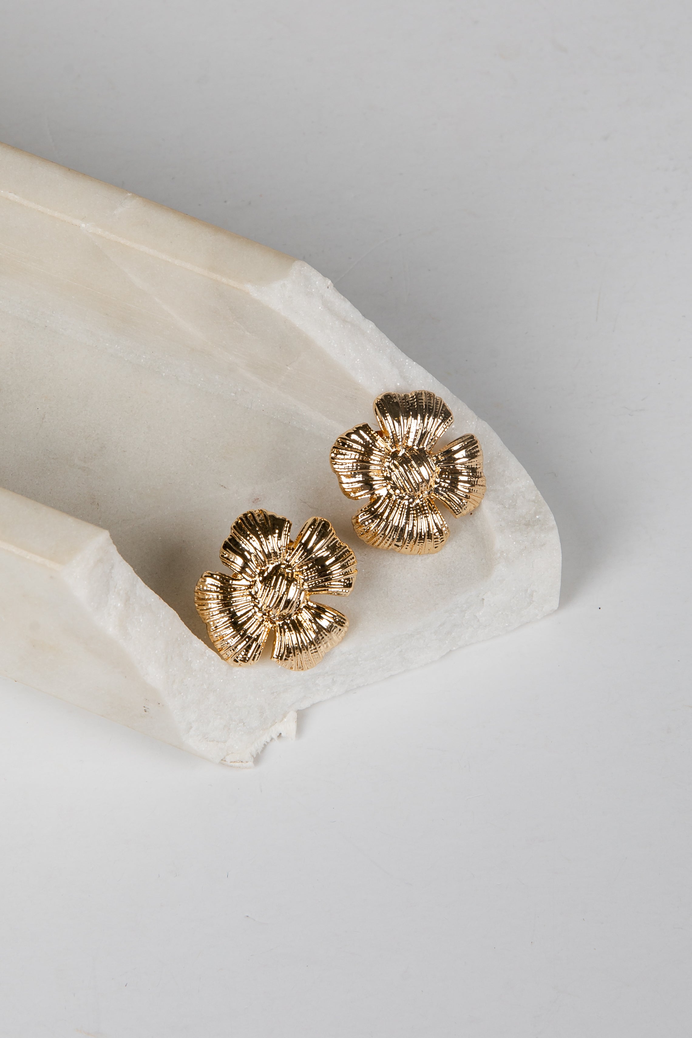 A pair of textured gold flower-shaped stud earrings resting on a white marble tray against a light gray background.