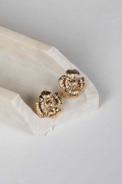 A pair of textured gold flower-shaped stud earrings resting on a white marble tray against a light gray background.