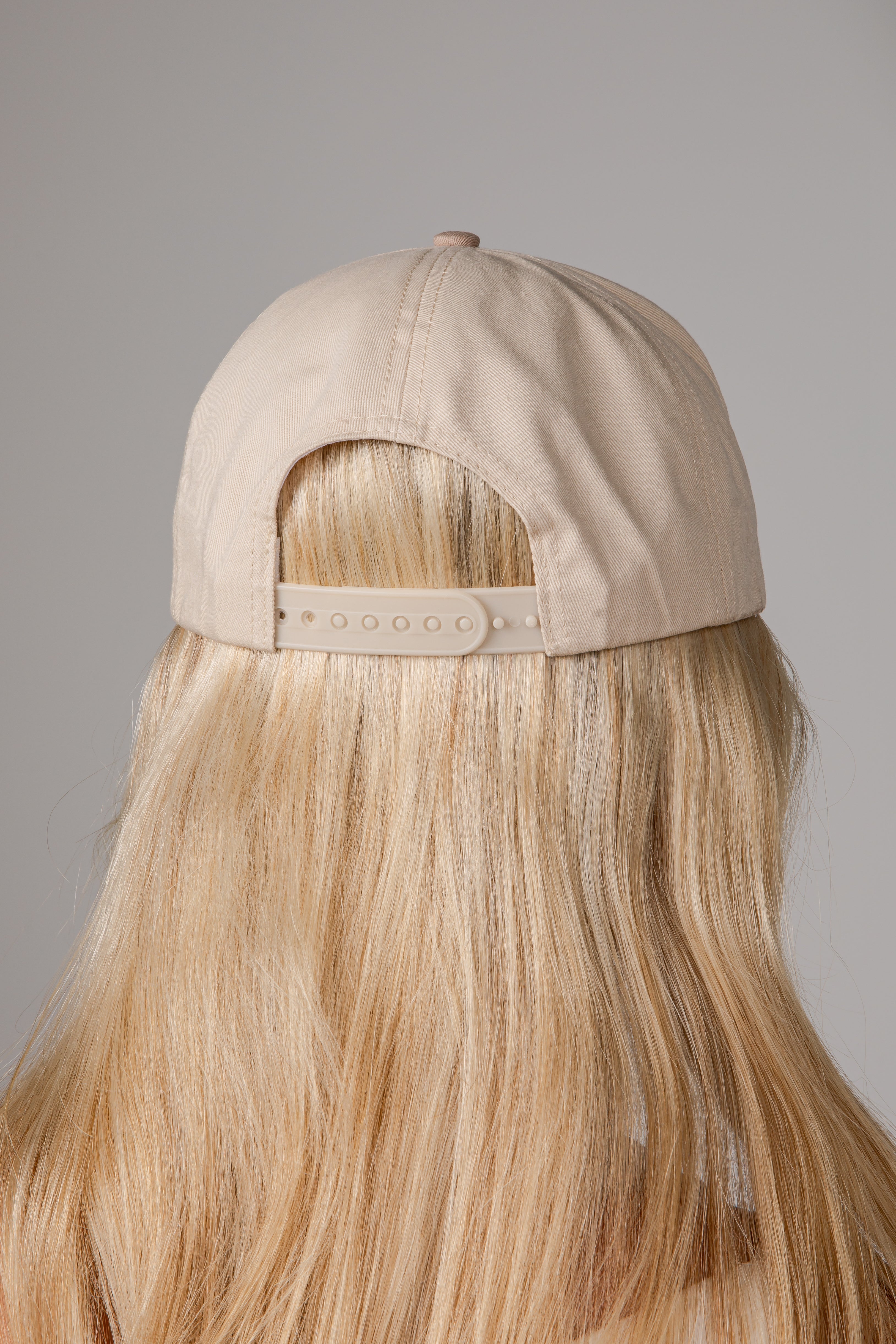 Back view of a beige baseball cap worn by a model with long blonde hair, shown against a neutral gray background.