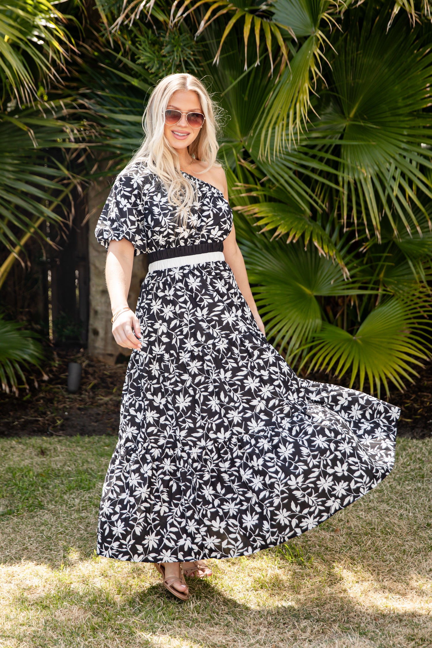 Woman in a black and white floral dress standing in front of green foliage.