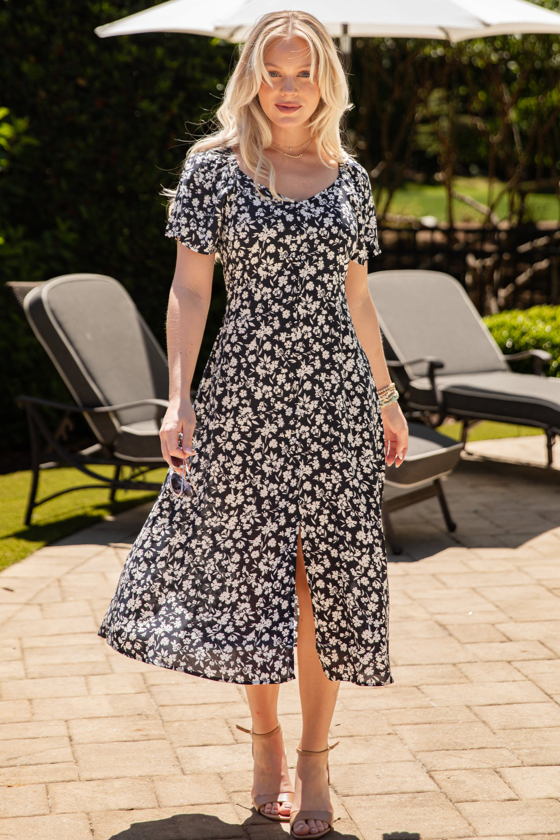 Woman in a floral dress standing outdoors on a patio with chairs and an umbrella in the background.