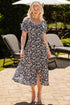 Woman in a floral dress standing outdoors on a patio with chairs and an umbrella in the background.