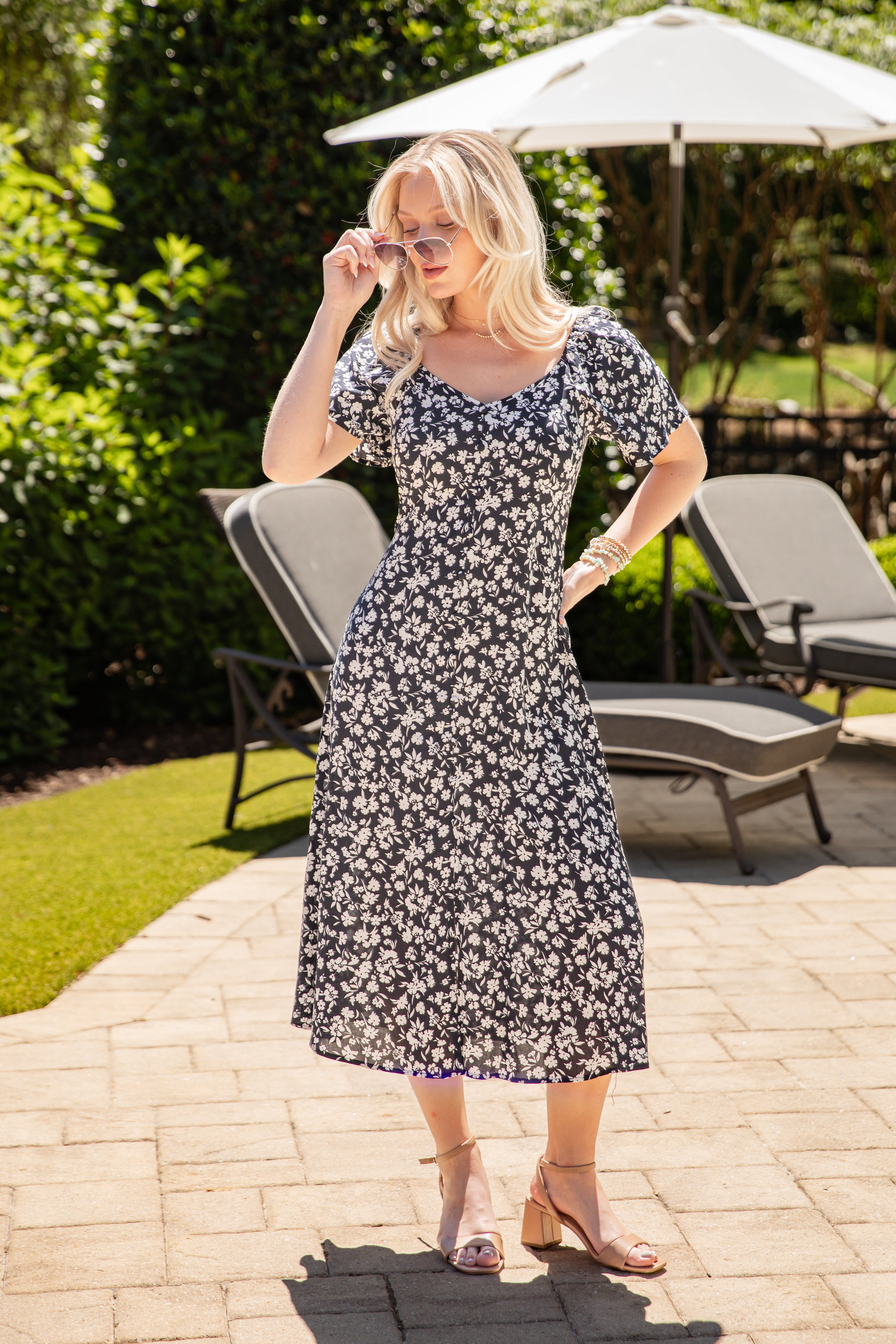 Woman in a floral dress standing outdoors on a patio with chairs and an umbrella in the background.
