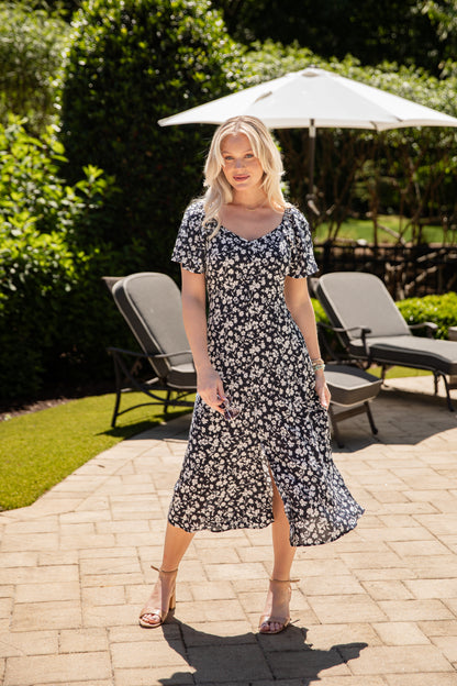 Woman in a floral dress standing outdoors on a patio with greenery and chairs in the background.