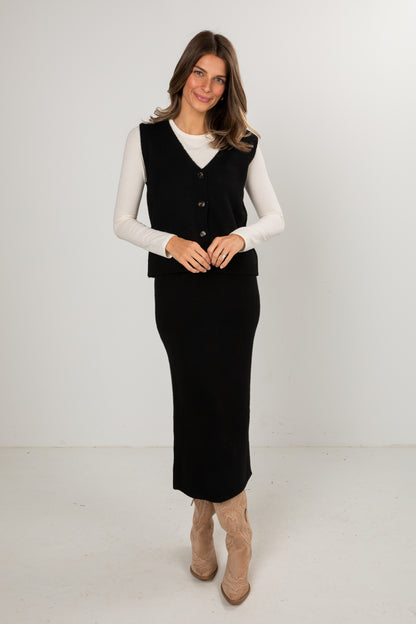 Woman wearing a black knit midi skirt and black buttoned sweater vest layered over a long-sleeve white top, styled with tan western boots, standing in a studio setting.
