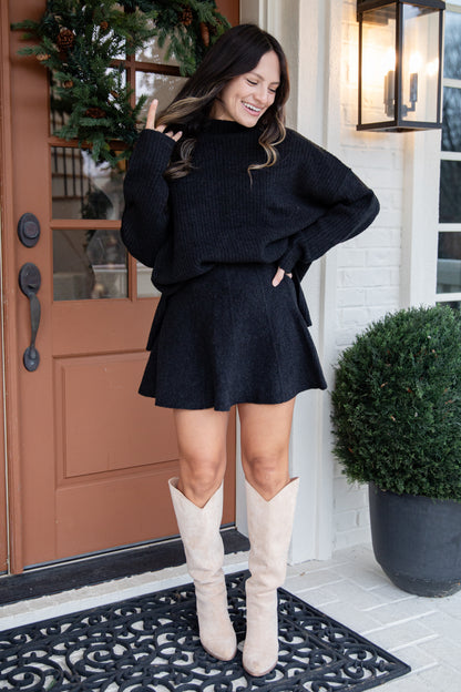 Woman in a black outfit with beige boots standing in front of a door.
