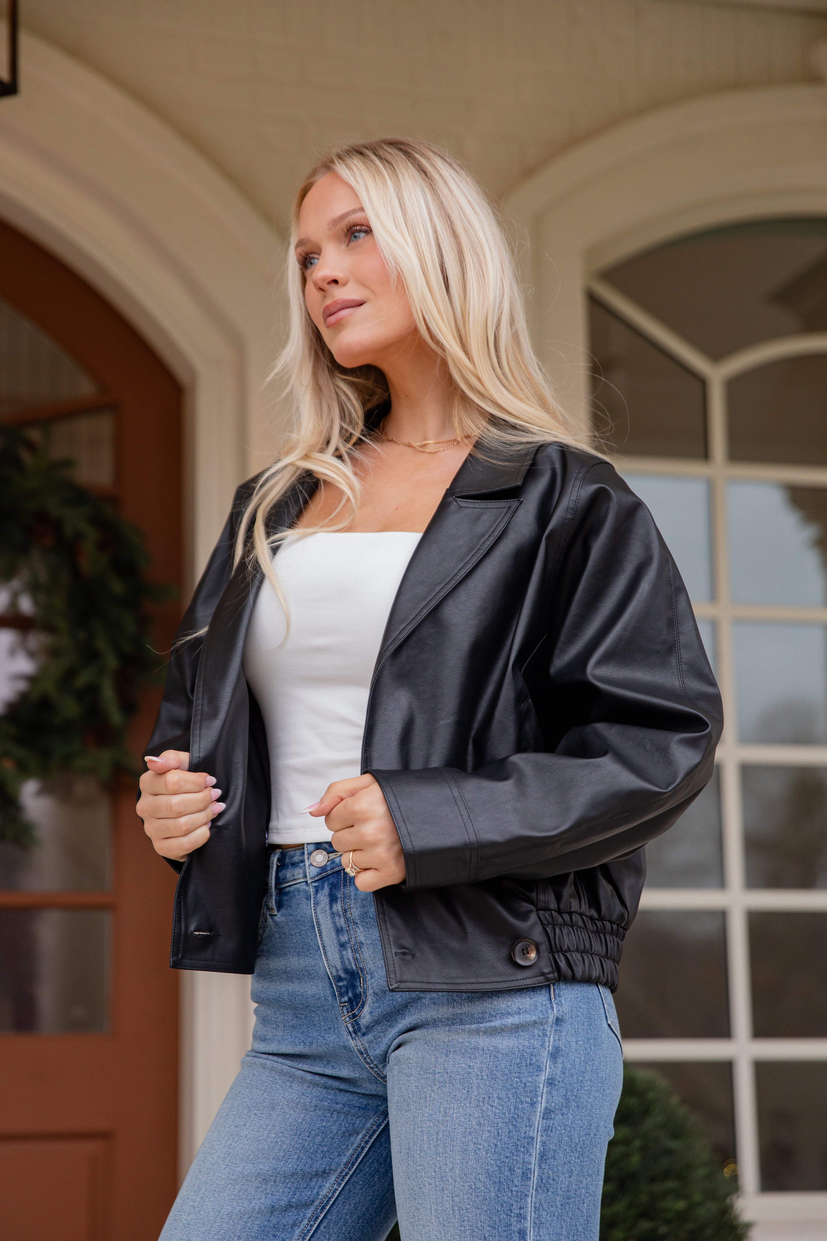 Woman wearing a black jacket over a white top and blue jeans, standing in front of a building entrance.