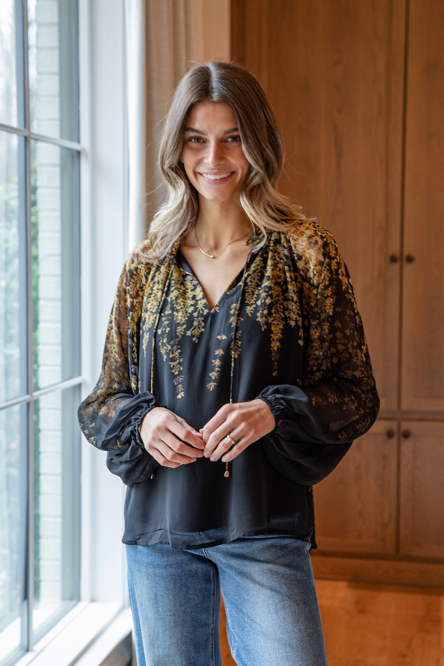Woman standing by a window wearing a patterned blouse and jeans.