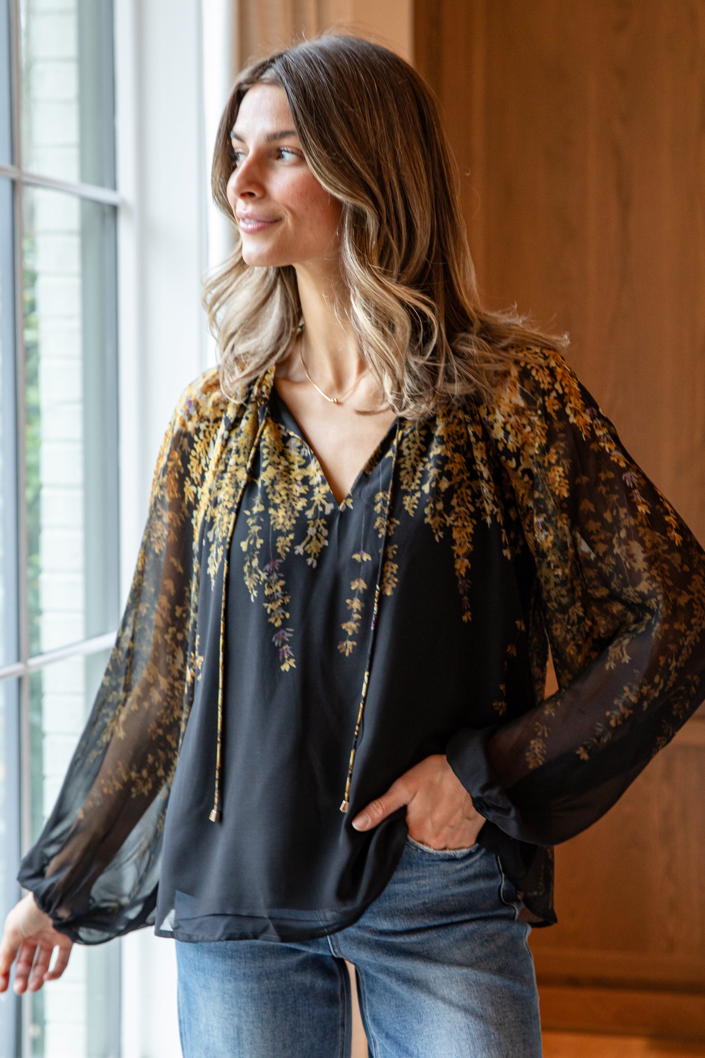 Woman wearing a black blouse with sheer sleeves and floral scarf, standing by a window.