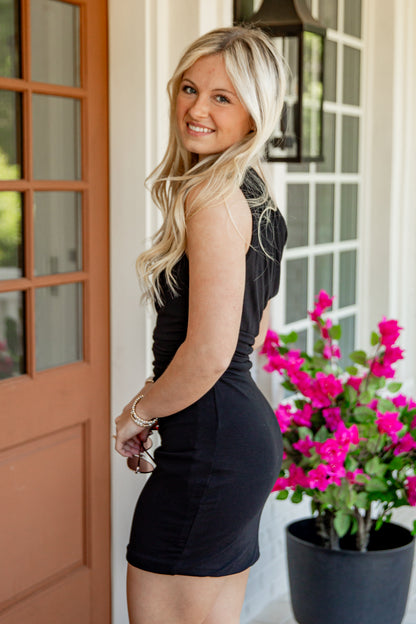 A blonde woman posing on a porch in a sleeveless black bodycon mini dress, with a brown door on the left and a potted plant with bright pink flowers on the right.