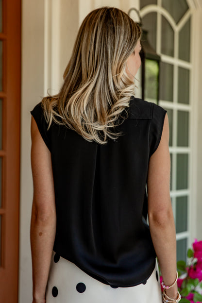 A woman viewed from behind wearing a sleeveless black top and a white skirt with black polka dots, standing in front of a white arched window.