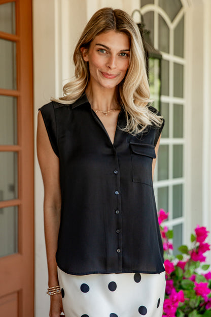 A woman wearing a sleeveless black button-up shirt with a chest pocket, paired with a white skirt with black polka dots, standing in front of a house porch with pink flowers.