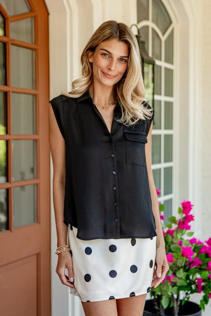 A woman stands on a porch wearing a sleeveless black button-front blouse and a white mini skirt with large black polka dots. The background shows a brown wooden door, white arched windows, and pink flowers.