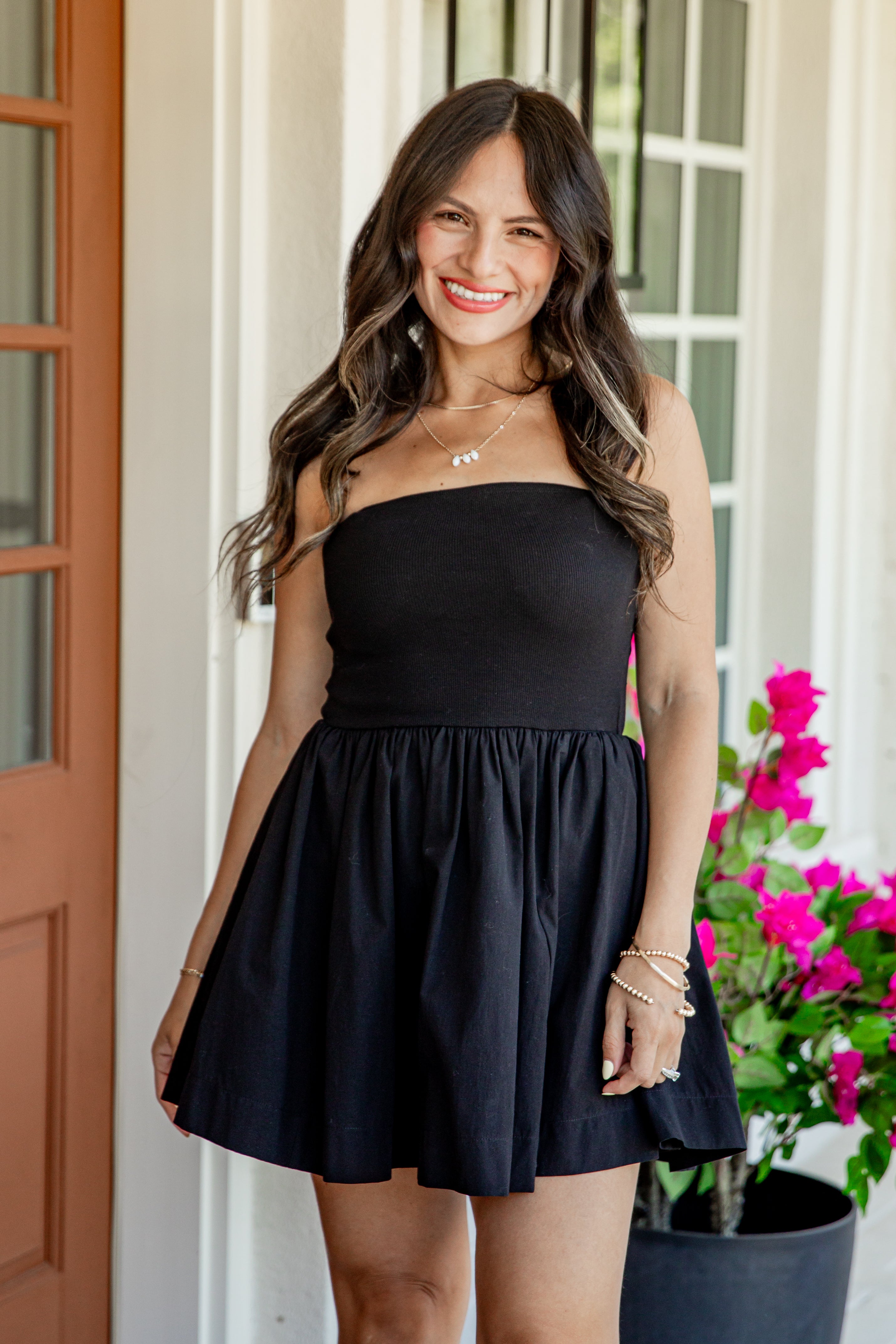 A woman modeling a black strapless mini dress with a gathered waist and flared skirt, standing on a porch with a potted plant of bright pink flowers in the background.