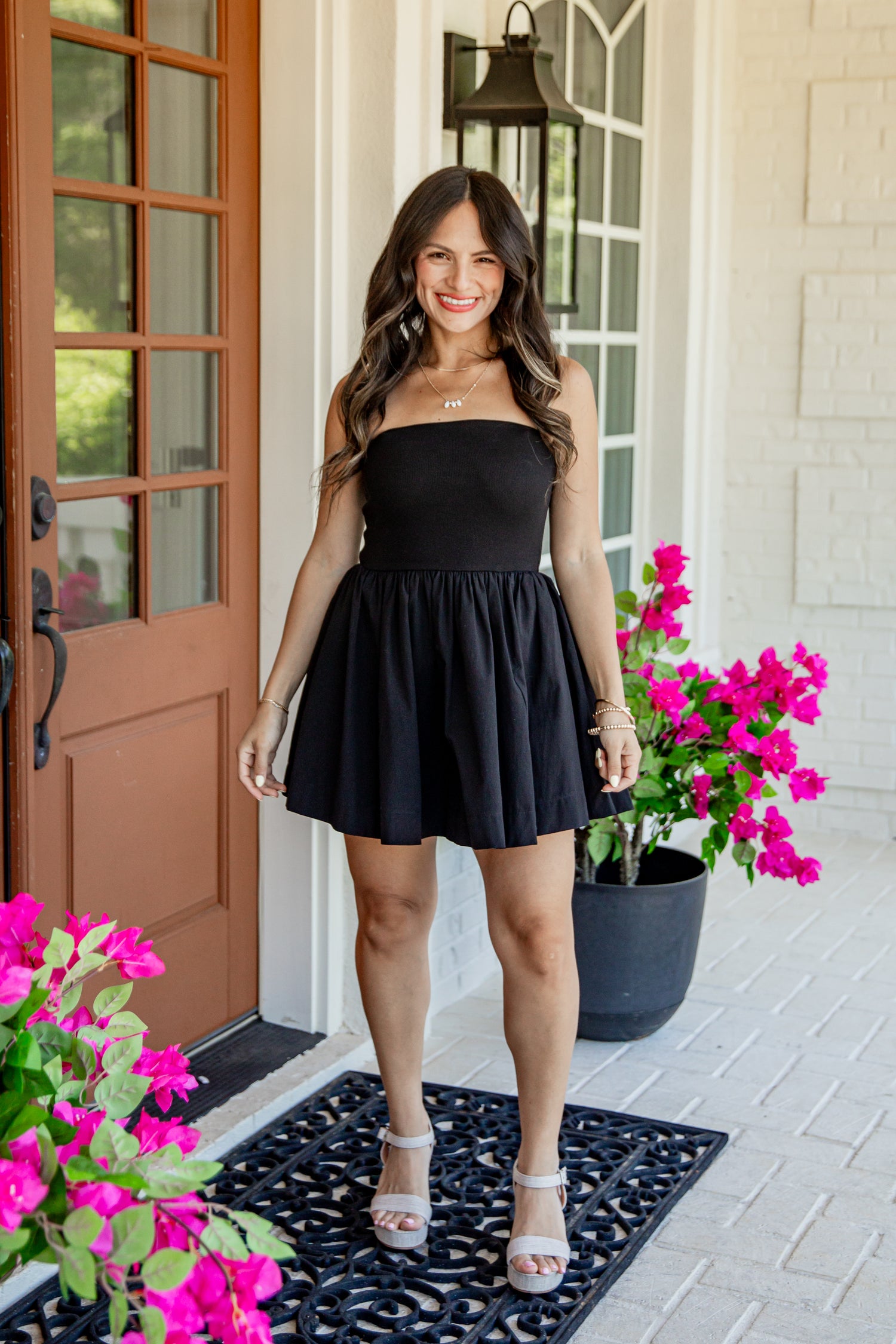 A woman stands on a front porch wearing a strapless black mini dress with a flared skirt and silver heeled sandals. Bright pink flowers and a brown door are visible in the background.