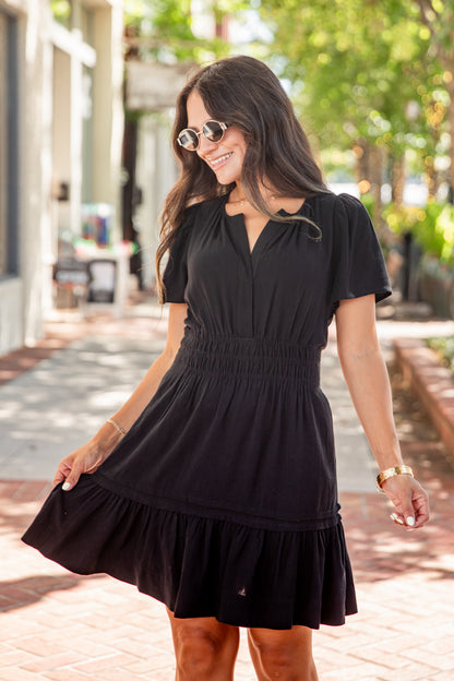 A woman wearing a black short-sleeve mini dress with a smocked waist and tiered hem, standing outdoors on a brick walkway with trees in the background.