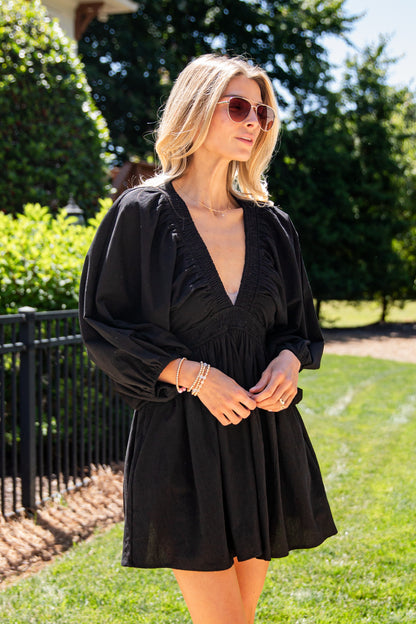A woman wearing a black long-sleeve mini dress with a deep V-neckline and gathered sleeves, standing outdoors on a grassy lawn with trees and a fence in the background.