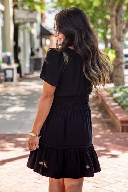 A woman with long dark hair is shown from behind wearing a black short-sleeve dress with a smocked waist and tiered skirt, standing on a brick sidewalk outdoors.