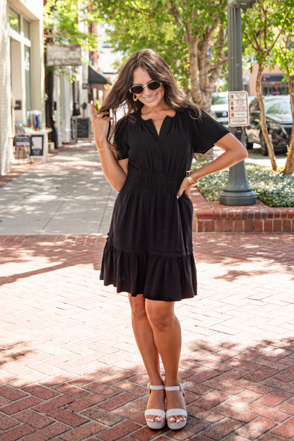 A woman wearing a black tiered mini dress with short sleeves and sunglasses, standing on a brick sidewalk in an outdoor urban setting.