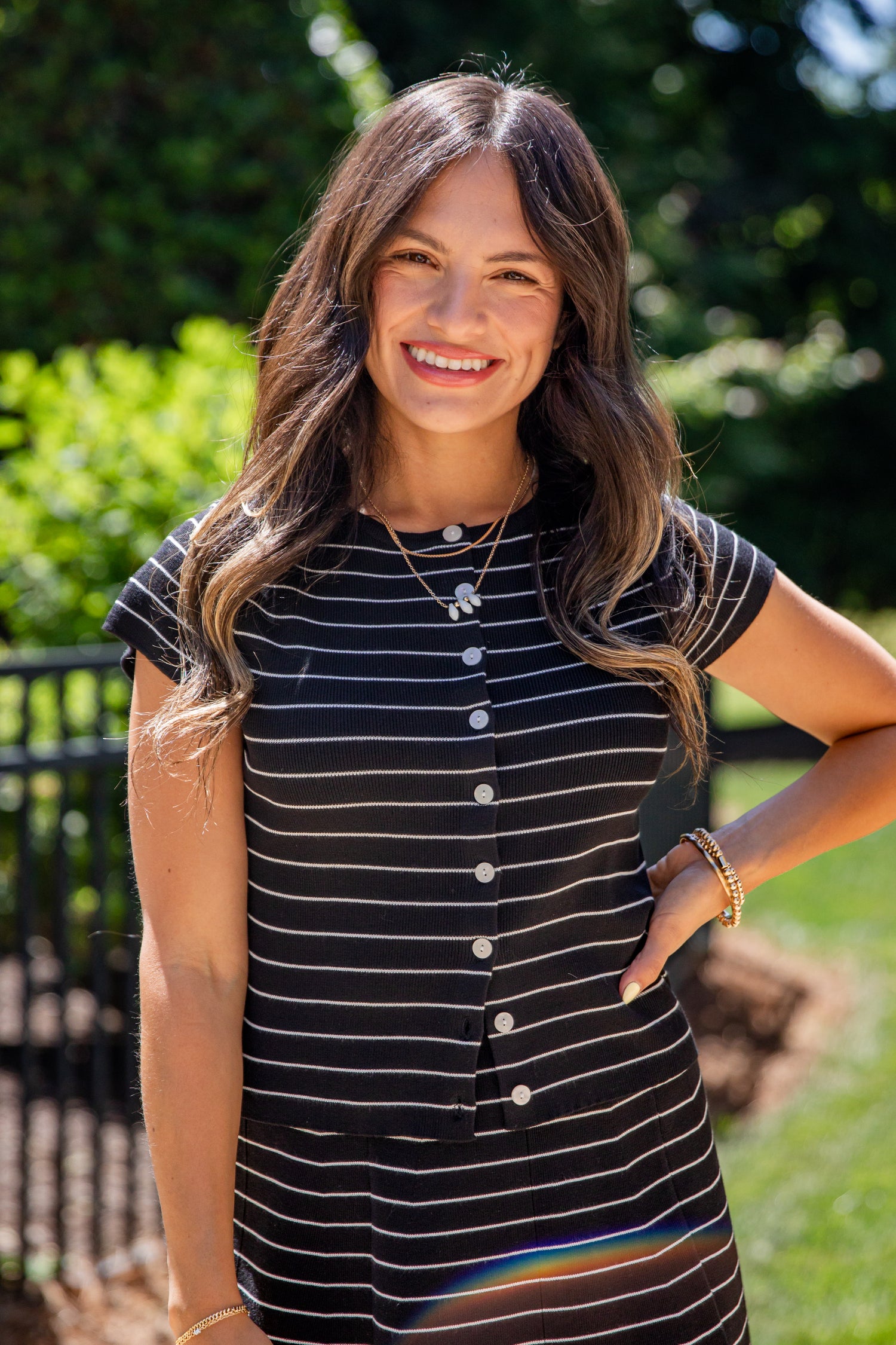 A woman smiling outdoors wearing a short-sleeve black and white striped button-front top , with green foliage and a fence in the background.