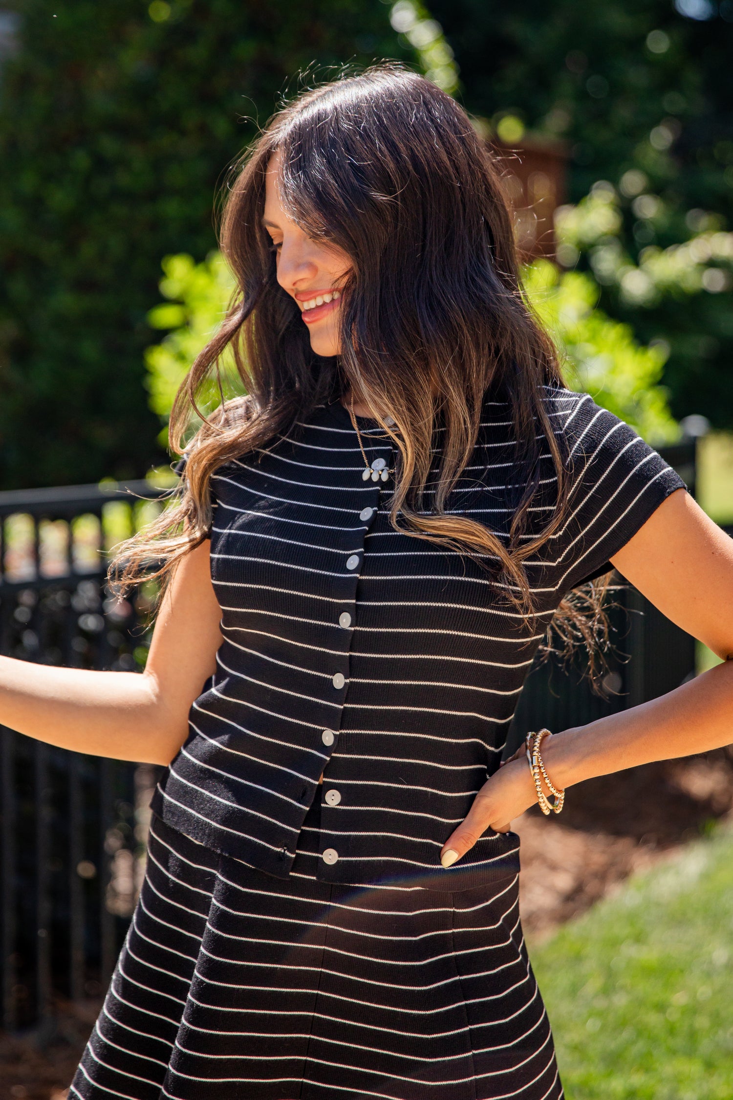 A woman wearing a black and white striped short-sleeve button-up dress, standing outdoors in a sunny garden setting.