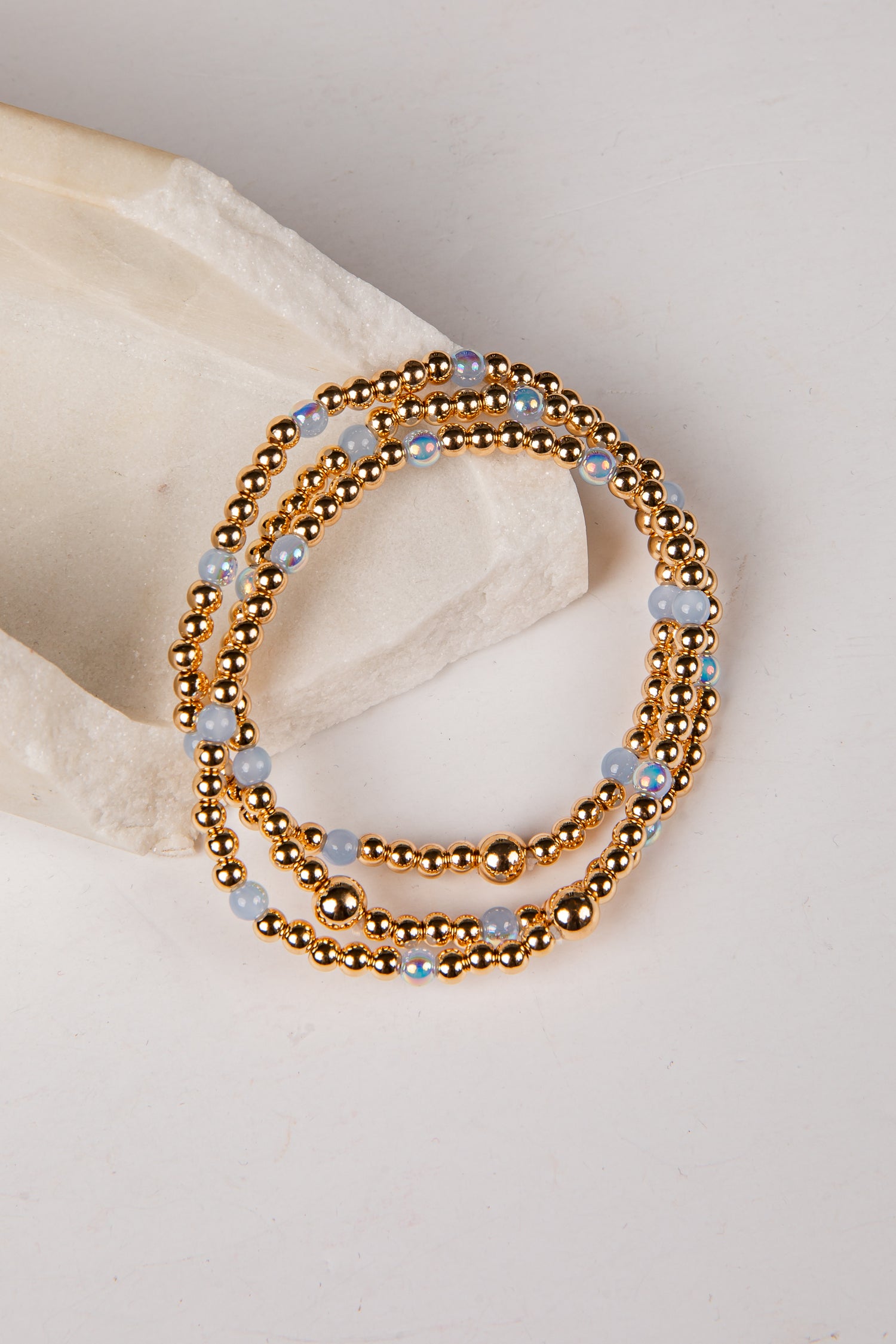 A stack of three beaded stretch bracelets with metallic gold beads and light blue faceted beads, displayed on a neutral cream-colored stone prop against a light background.