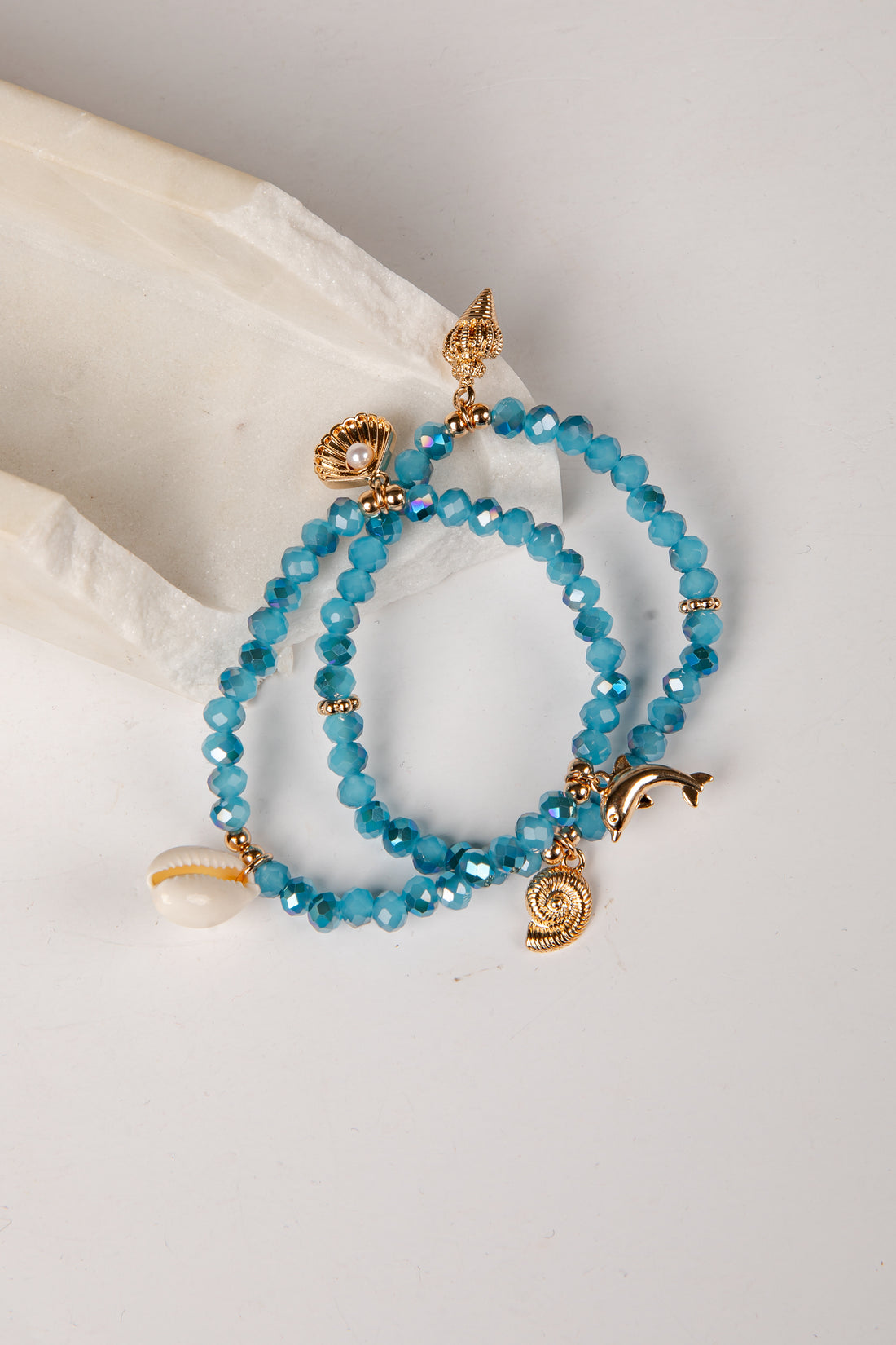 A pair of beaded bracelets with faceted turquoise-blue beads and gold-tone charms, including a dolphin, spiral shell, and small gold spacer beads, displayed on a white fabric prop against a white background.