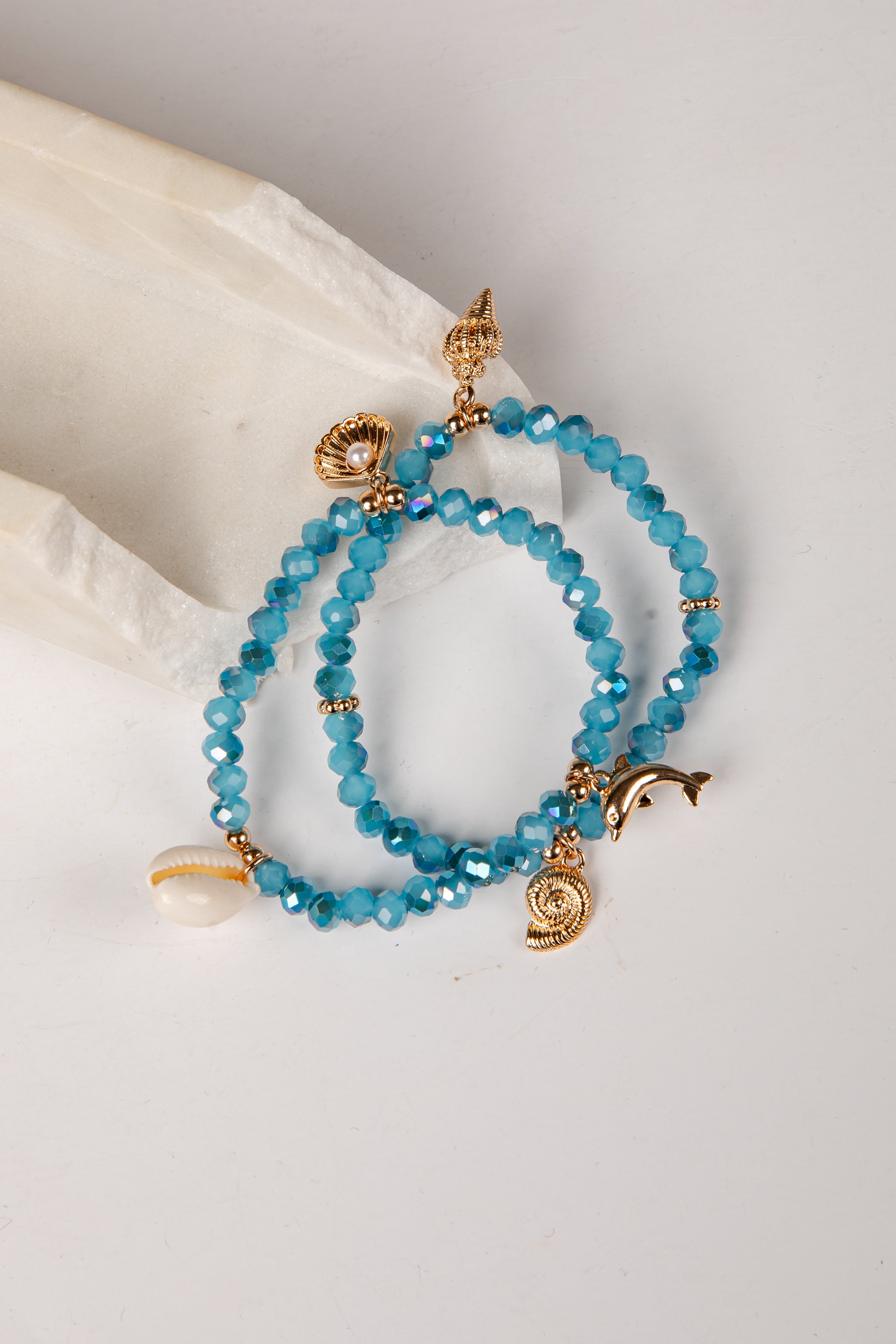 A pair of beaded bracelets with faceted turquoise-blue beads and gold-tone charms, including a dolphin, spiral shell, and small gold spacer beads, displayed on a white fabric prop against a white background.