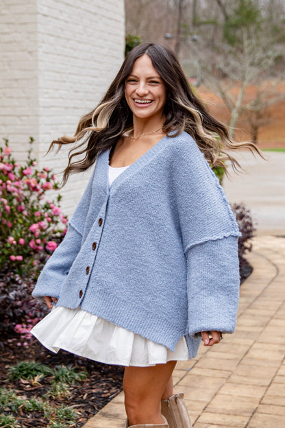 Woman wearing a blue cardigan over a white dress, standing outdoors with flowers and a building in the background.