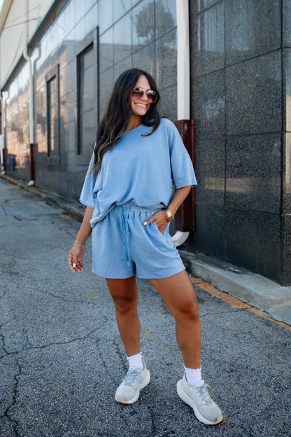 A woman wearing a light blue oversized t-shirt and matching drawstring shorts, styled with white socks and light gray sneakers, standing on a paved street next to a dark gray building.