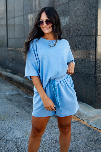 A woman wearing a light blue short-sleeve t-shirt and matching drawstring shorts, standing outdoors against a dark stone wall.