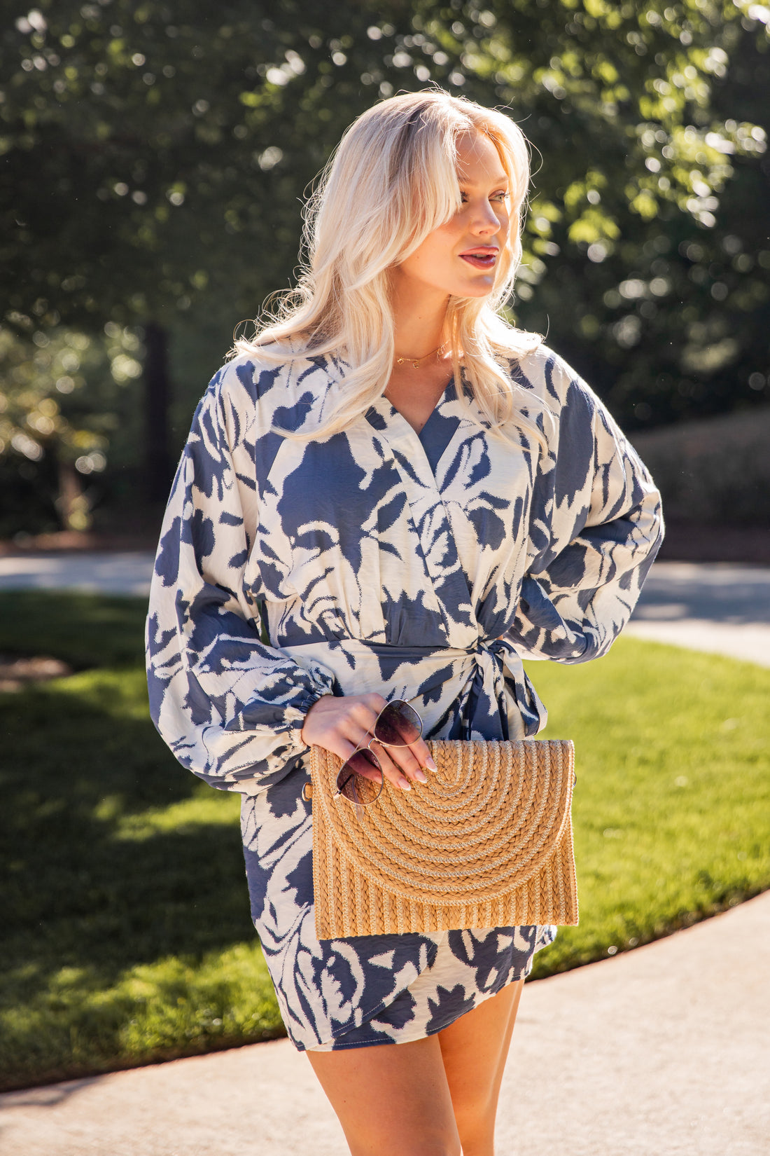 Model wearing a blue floral print wrap mini dress with long sleeves and a waist tie, styled with a woven clutch outdoors.