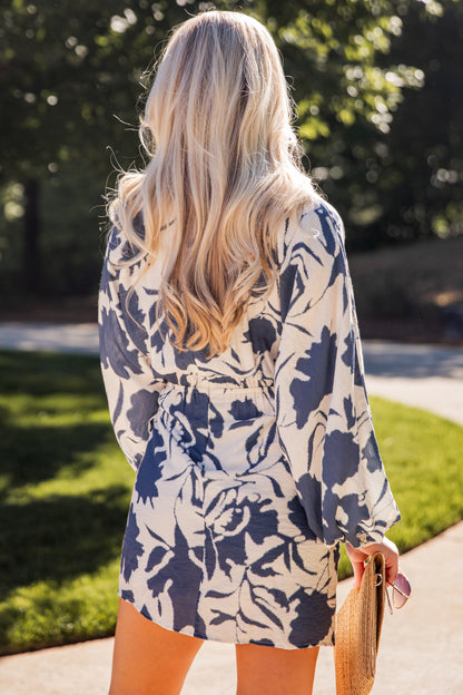 Model wearing a blue floral print wrap mini dress with long sleeves and a waist tie, styled with a woven clutch outdoors.