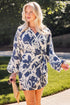 Woman wearing a blue and ivory floral print wrap mini dress with long balloon sleeves and a waist tie, holding a woven clutch while walking outdoors in the sunshine. The relaxed silhouette and bold botanical print create an effortless, elevated look perfect for summer events and vacations.