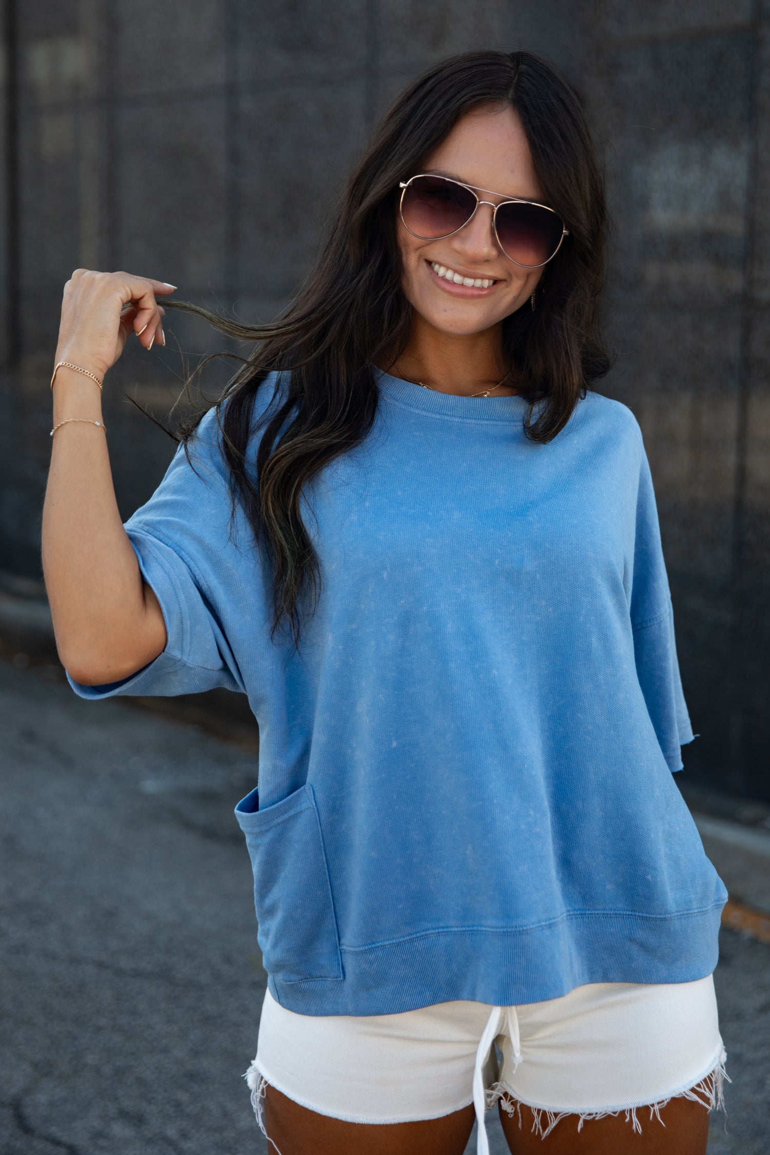 A woman wearing a light blue oversized crewneck sweatshirt and white frayed denim shorts, posing outdoors against a dark gray concrete wall.